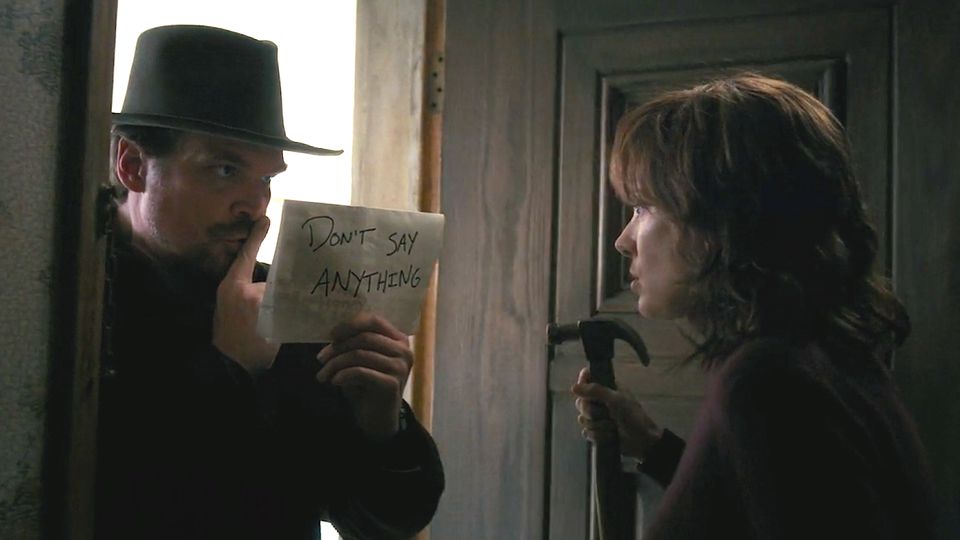 A man in a wide-brimmed hat stands in a doorway with a finger to his lips and a sign that reads Don't Say Anything. A woman stands in front of him holding a hammer.