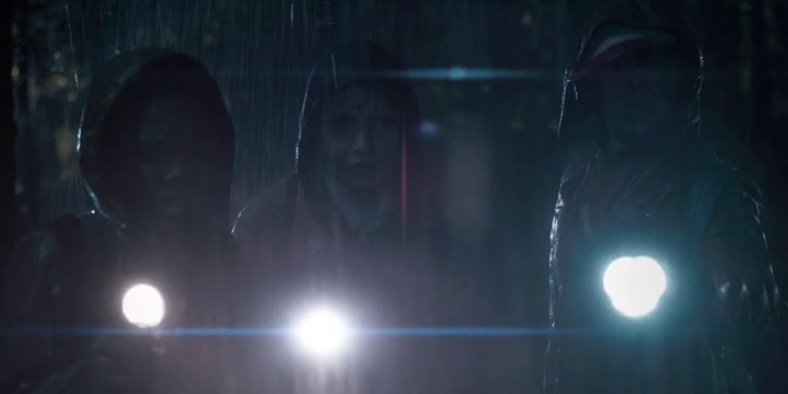 Three boys in raincoats stand in the dark and pouring rain, each holding a flashlight.
