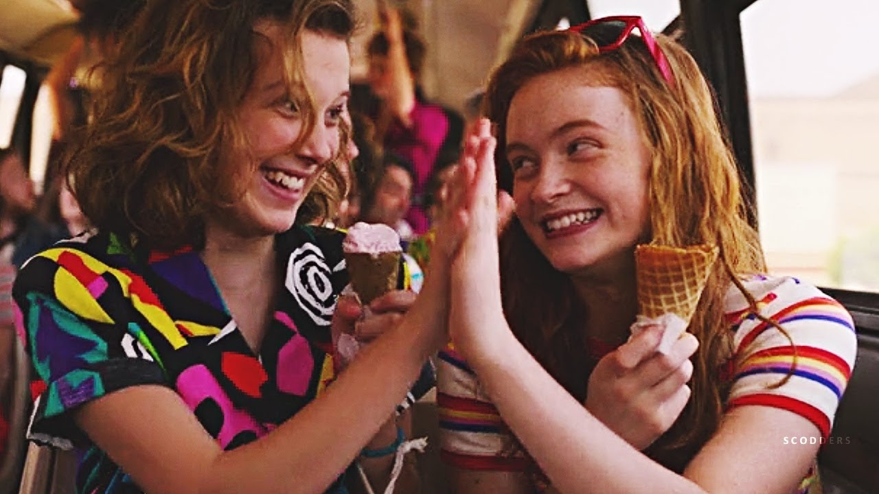 Two young girls wearing bright colors and holding ice cream cones smile at each other with a high five.