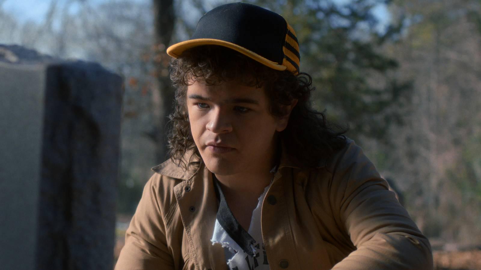 A teenage boy with curly hair and a baseball cap looks sadly at a tombstone.