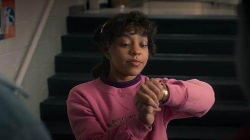 A teenage girl with curly black hair holds her arm up, checking the time on her watch. She is in a school hallway.