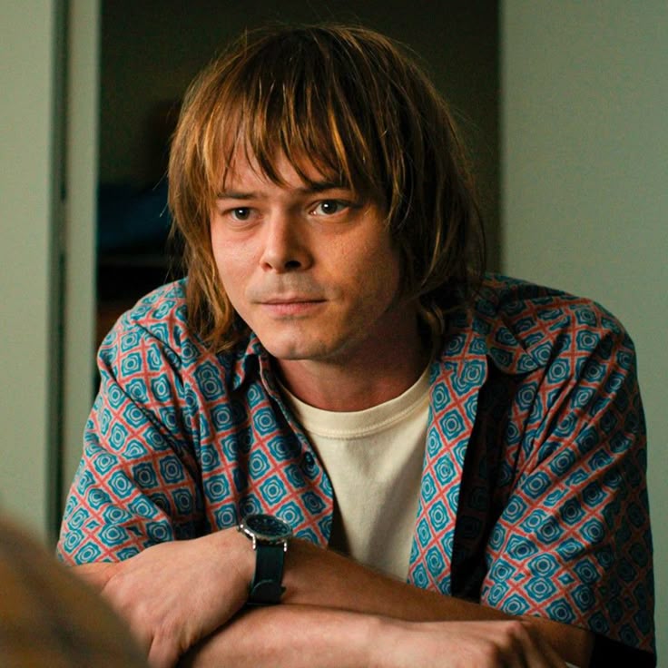 A young man with shaggy hair and a colorful unbuttoned shirt talks to someone with a kind and understanding expression.