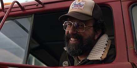 A man with a beard, glasses, and a baseball cap leans out the window of the truck he is driving with a grin on his face.
