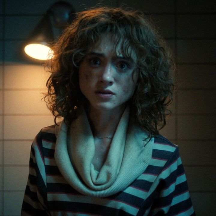 A young woman with shoulder-length hair stares forward in fear and shock. Behind her is a harsh light fixture on a plain white bricked wall.