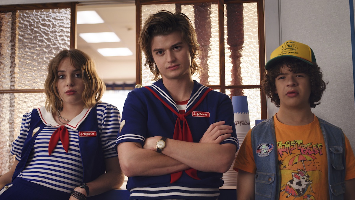 Two older teenagers, a girl with a nametag that says Robin and a boy with one that says Steve, lean against a counter wearing nautical themed blue, white, and red work outfits. A casually dressed younger teenage boy stands next to them.