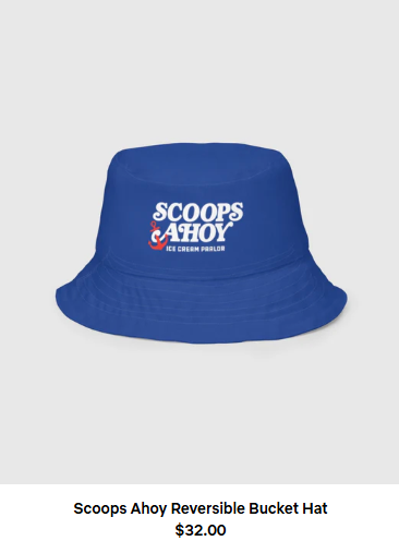 A blue bucket hat with a white logo that says Scoops Ahoy Ice Cream Parlor. Words beneath it say Scoops Ahoy Reversible Bucket Hat $32
