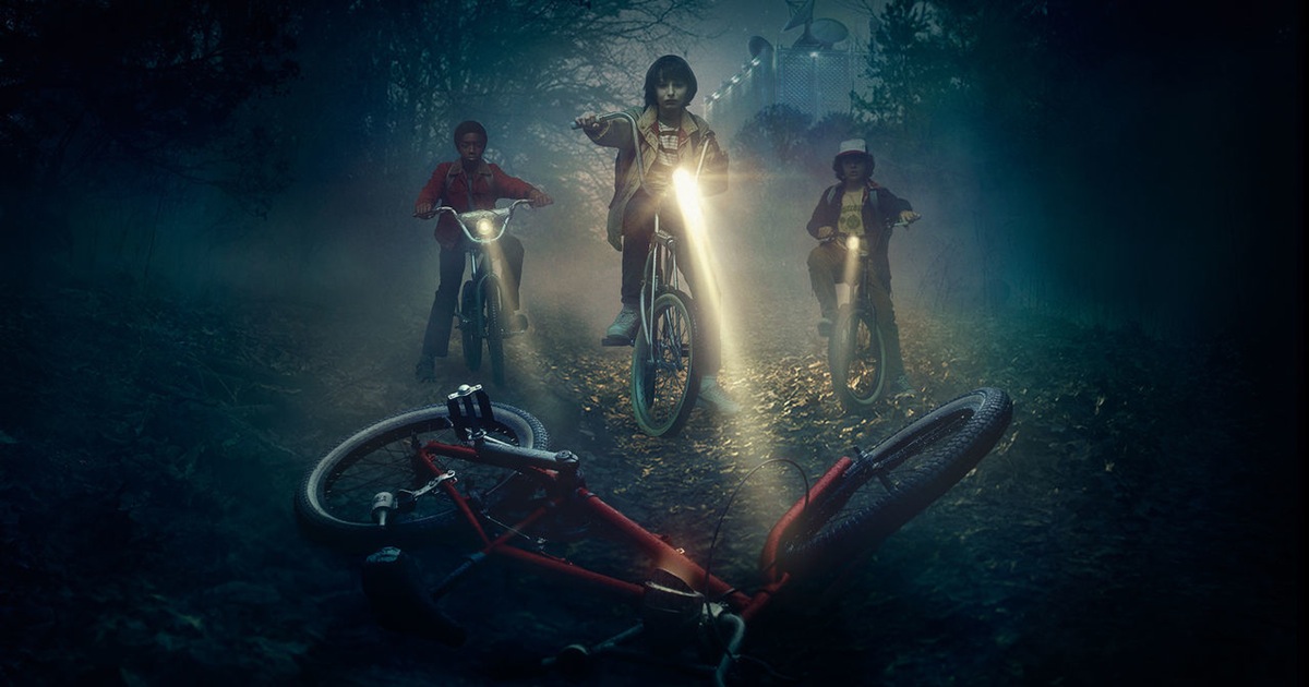 Three young boys on bikes look down solumnly at an abandoned bike. They appear to be in a dark forest and the fog is thick, catching the light beams from their flashlights.
