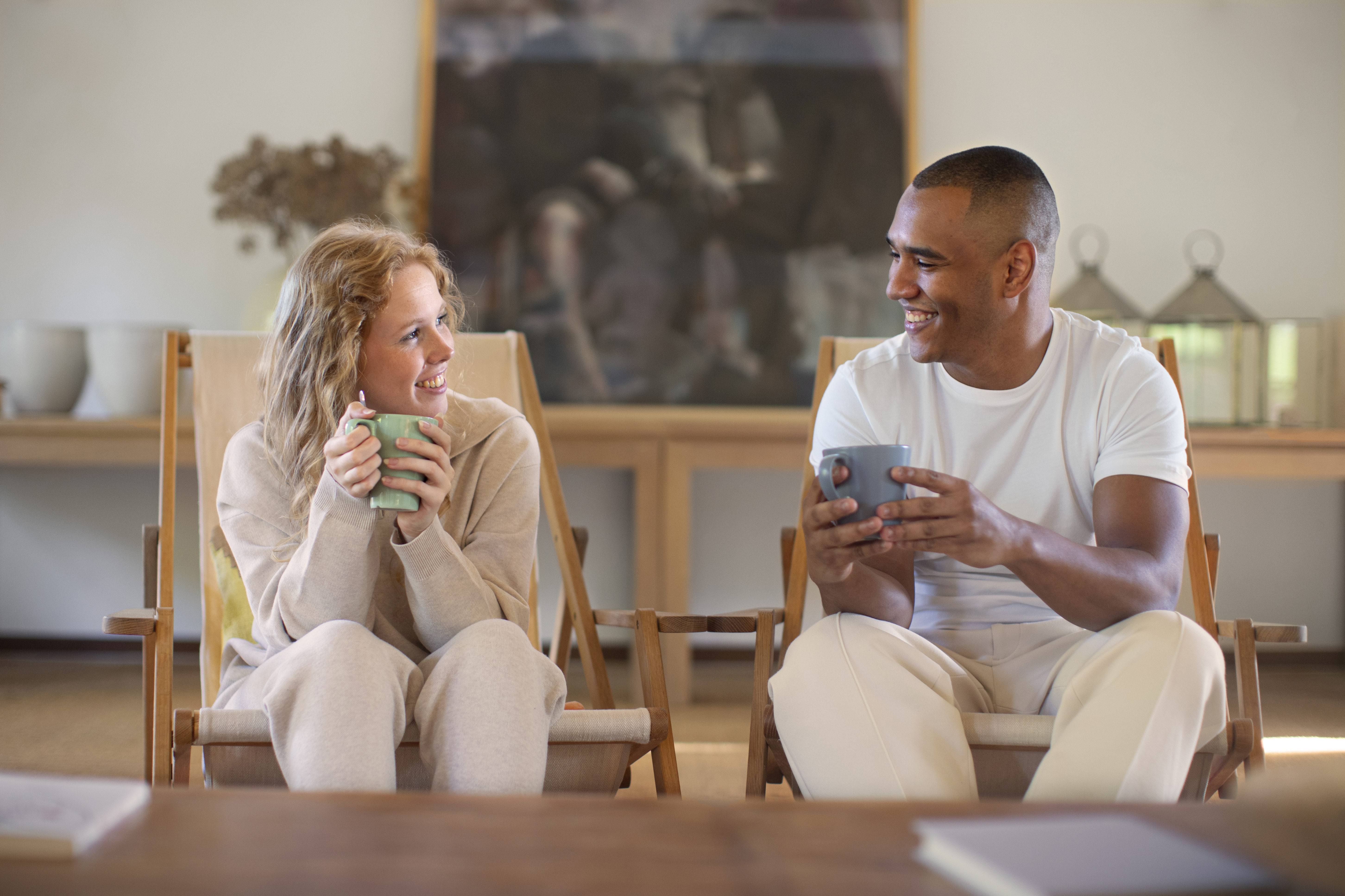 two people smiling at each other while they sit in adjacent chairs and each hold a mug in their hands, the background shows an interior with neutral colored furniture and decor