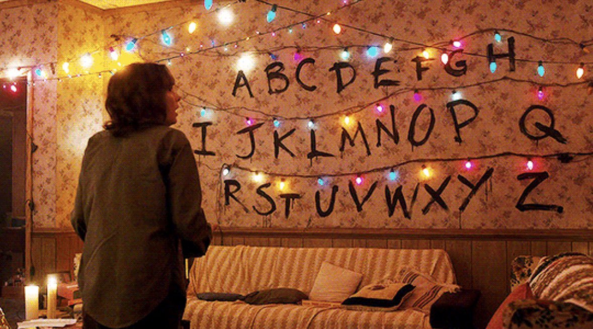 A woman stands in front of a wall that has the alphabet messily painted on it. A strand of Christmas lights is strung across it so that an illuminated light is over each of the letters.