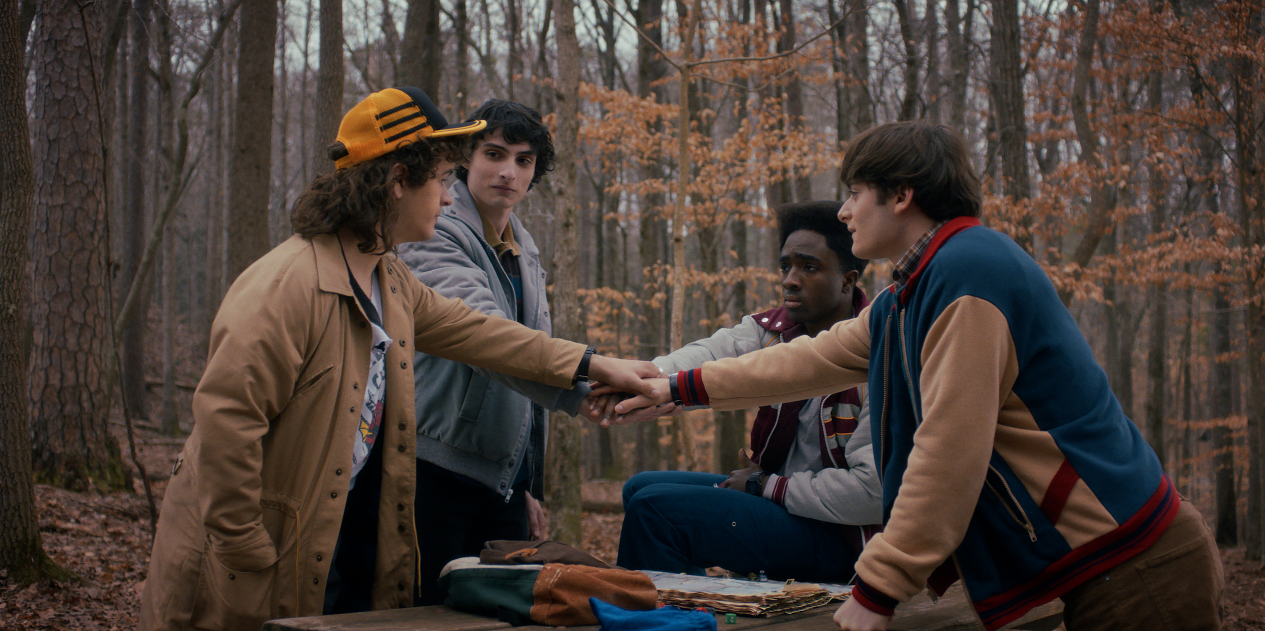 Four teenage boys stand around a picnic table in the woods. They each reach forward to stack their hands in the middle.