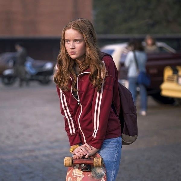 A young red-haired girl holds a skateboard in front of a school. 