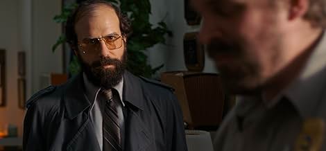 A man in a suit and tinted glasses waits to speak to a police chief.