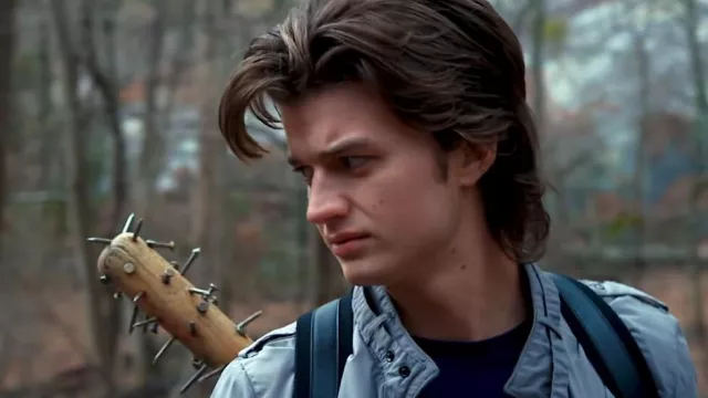 A teenage boy with nice hair carries a bat with nails pounded into the end in a backpack while he looks off to the side.