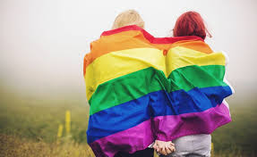 A pair of women stand close and hold hands as they wrap the pride flag around their shoulders 
