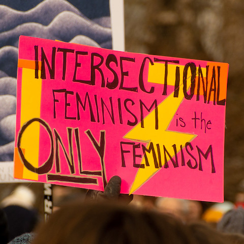 A pink, yellow, and orange sign held up over an out of focus group of people. It reads 'Intersectional feminism is the only feminism'