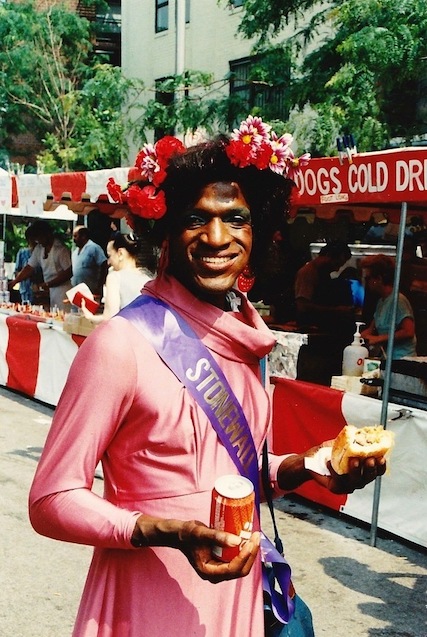 Marsha P. Johnson in makeup smiling at the camera. She is wearing a pink dress with a purple sash that says 'Stonewall' in silver lettering.