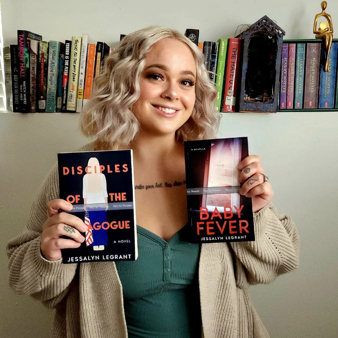 Photo of Jessalyn LeGrant smiling holding up two of her books 'Disciples of The Demagogue' and 'Baby Fever' with a plain wall and a shelf of books in the background