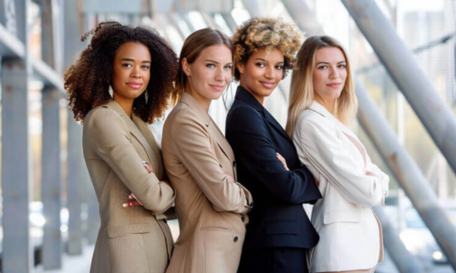 four ethnically diverse women with their arms crossed in front of them standing next to each other in varying colors of business suits smiling at the camera