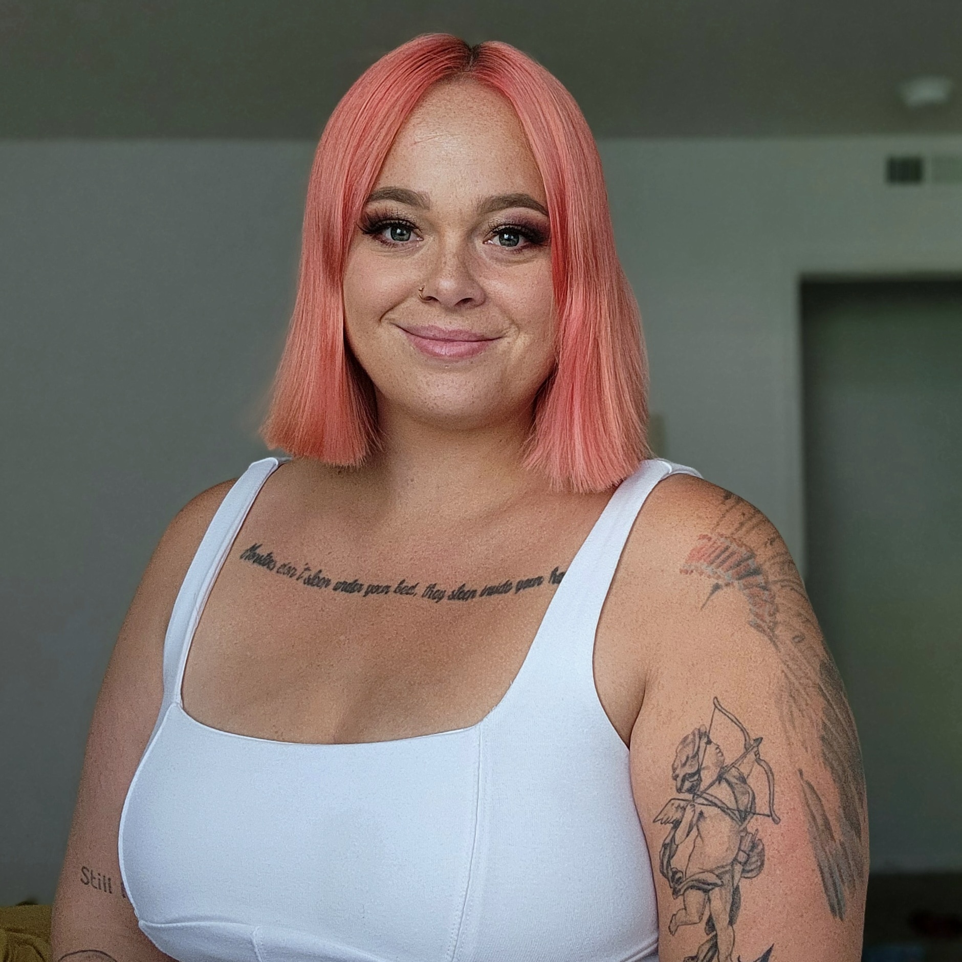 Photo of a woman with pink hair and wearing a white tank top. She has tattoos on her chest and arms. She is smiling.