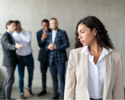 photo of a woman wearing a business suit is standing close to the camera while four men dressed in business suits are blurry in the background and are laughing and pointing and gossiping about the woman.