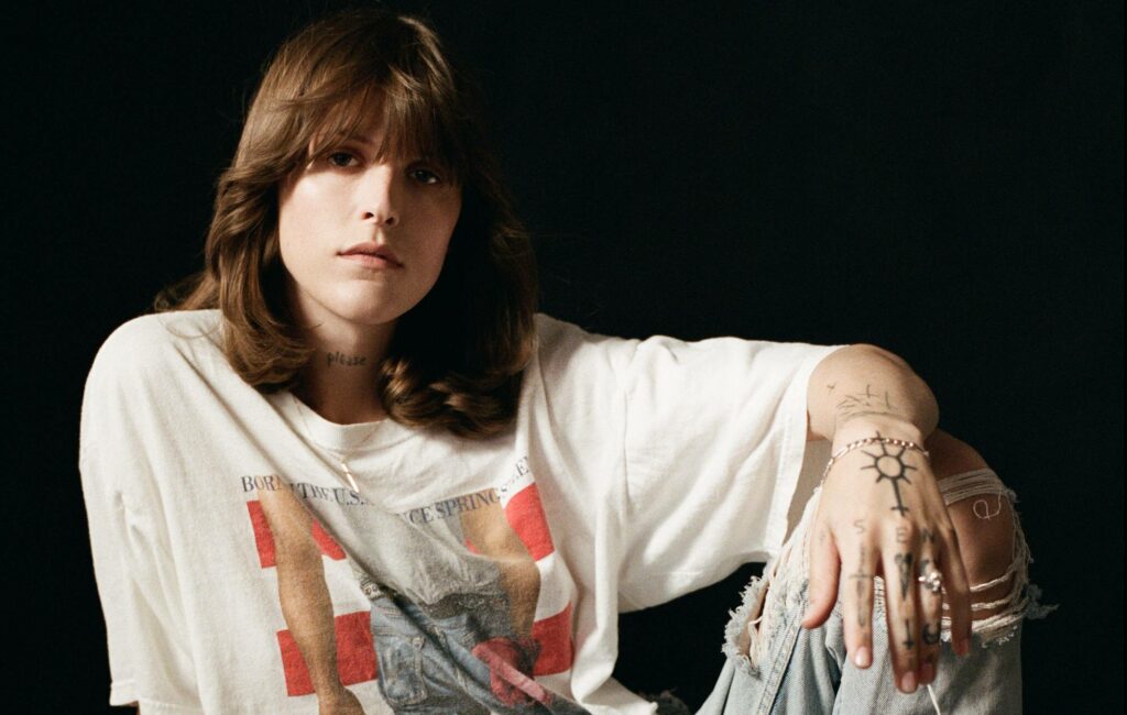 Ethel Cain sits up leaning on her arm with a shaggy wolf cut, wearing a Bruce Springsteen t-shirt in front of a black background