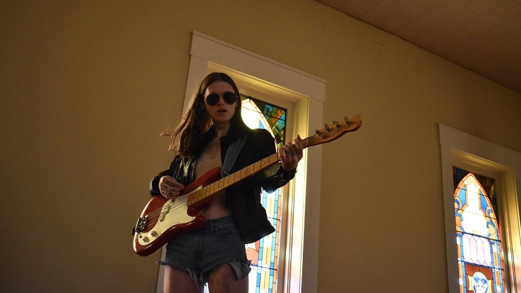 Ethel Cain plays the guitar in a chapel with stained glass windows behind her while wearing aviator sunglasses and a leather jacket