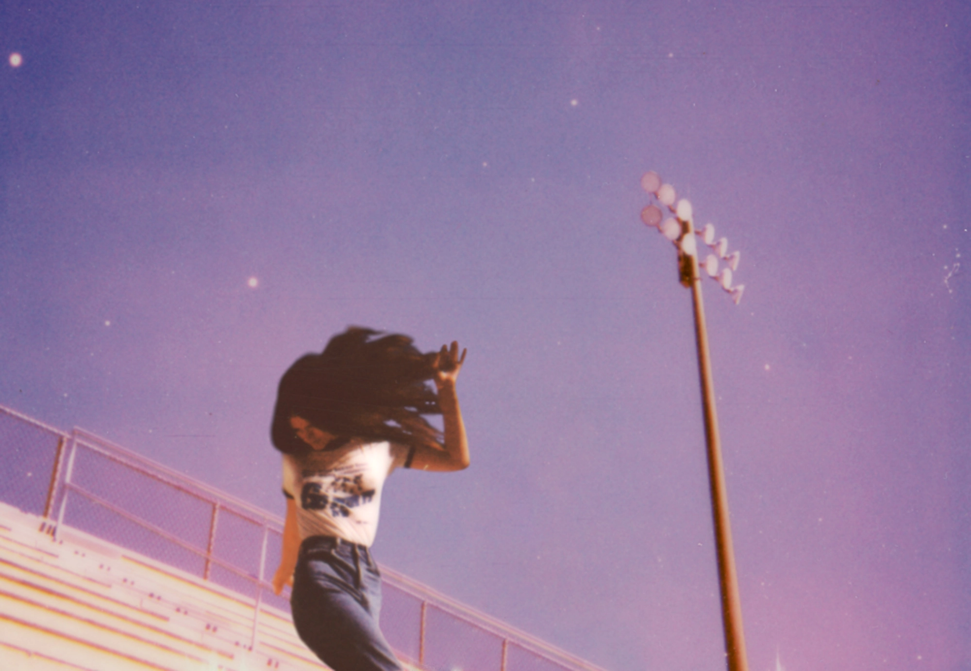 Ethel Cain dancing on the bleachers with a clear blue sky and a single floodlight behind her