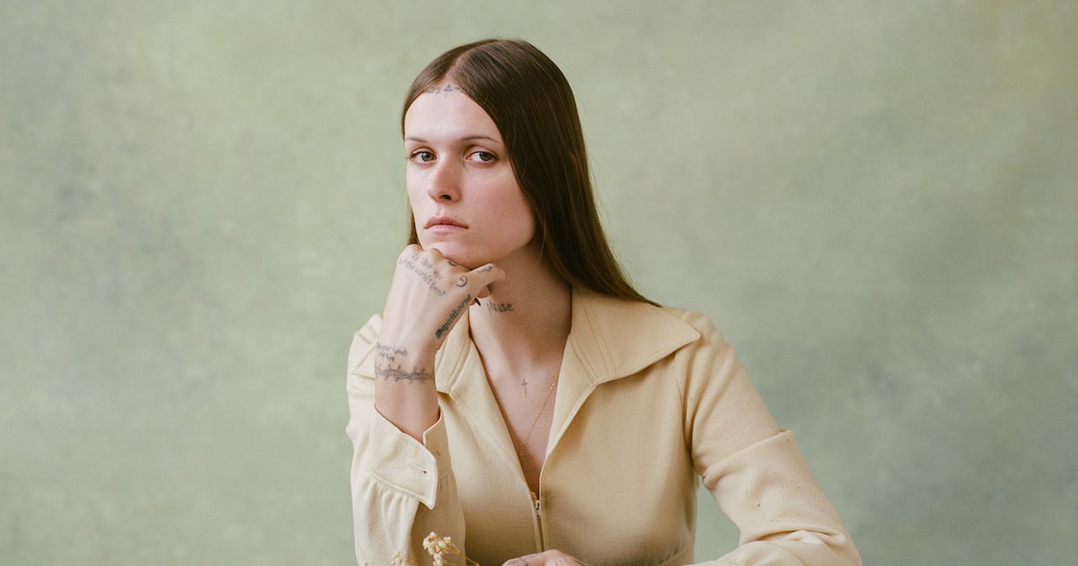 Ethel Cain posed with her hand on her chin looking at the camera. Dried Flowers in her hand with a grey wash background.