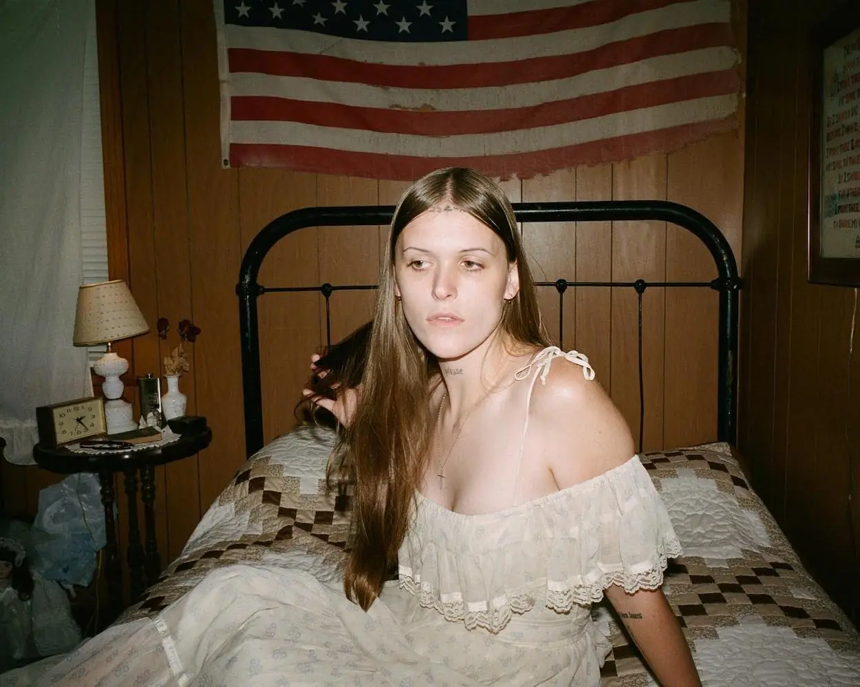 Ethel Cain wearing a low cut white dress on a twin size bed with metal bedframe with tattered American Flag on the wall behind her