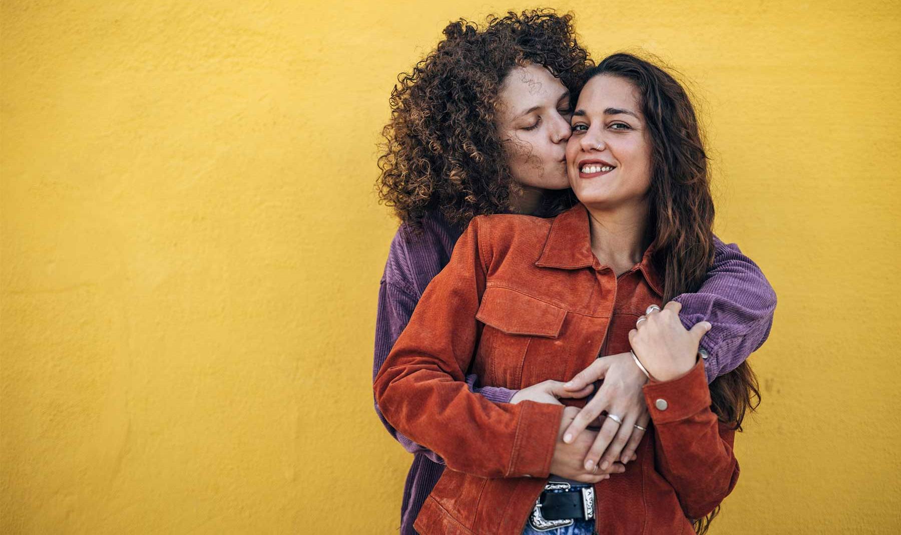 Two women embracing, one is hugging the other from behind while kissing her