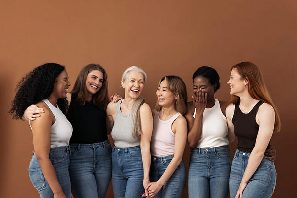 Six women of different age and color standing alongside each other either holding hands or having arms wrapped around each other while looking at one other and laughing