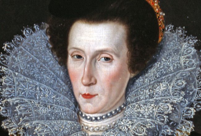 A painting depicts a portrait of a tudor-era, royal woman. She is wearing a wide, lacy collar and has a peaceful and subtle smile on her face. Her cheeks are rosy. She is wearing a pearl necklace. She has brown, curly hair that is tied back from her face.