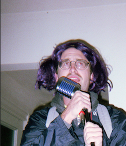 A picture depicts the low angle of a mustached man in the center of a dimly-lit room. He is holding an old, fifties-style microphone and singing or yelling into it with a determined expression on his face. He is wearing a cheap, polyester, purple wig that sticks out from his head at odd angles and large perscription glasses. He is wearing a blue bomber jacket and a backpack.