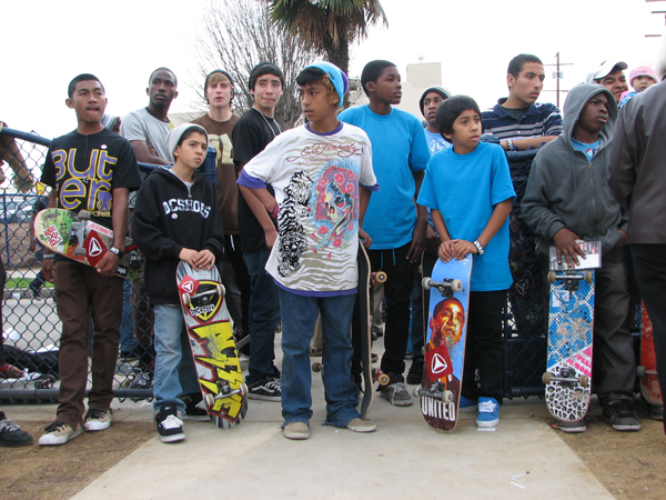 A group of teenaged-looking individuals stands together. They are all wearing casual clothes; T-shirts and beanies with loose jeans. A good majority of them are holding skateboards. Behind them is a visible palm tree.