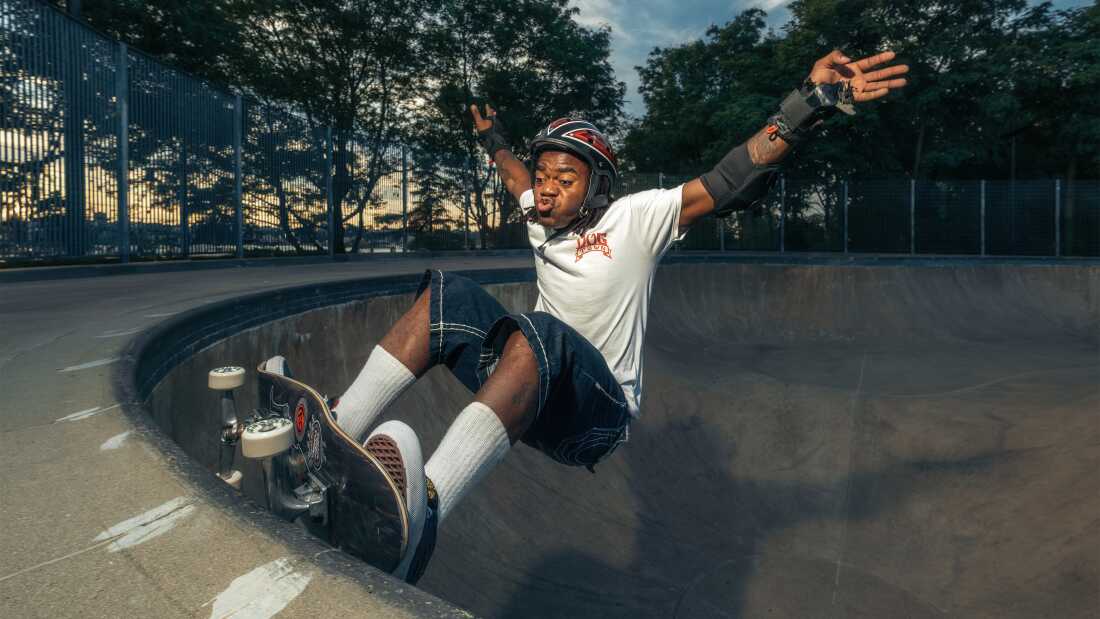 A young man is using a skateboard to skate in a half-pipe. He has a concentrated expression on his face as he flails his arms out to his sides and balances on the edge.