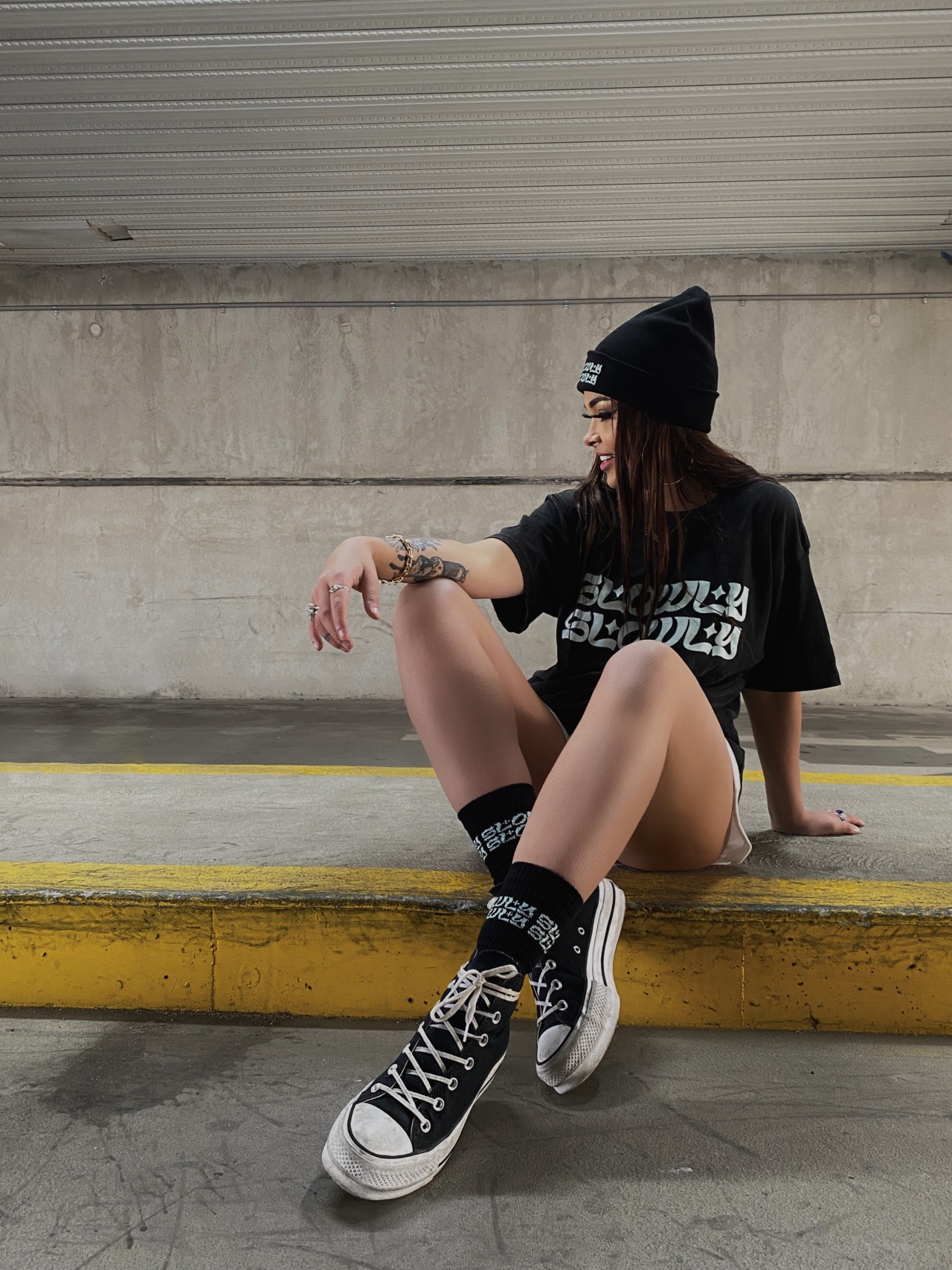 A young woman sits on the curb of an underground subway, looking to her left so her profile is visible. She is wearing a black beanie and shirt. She is also smiling.