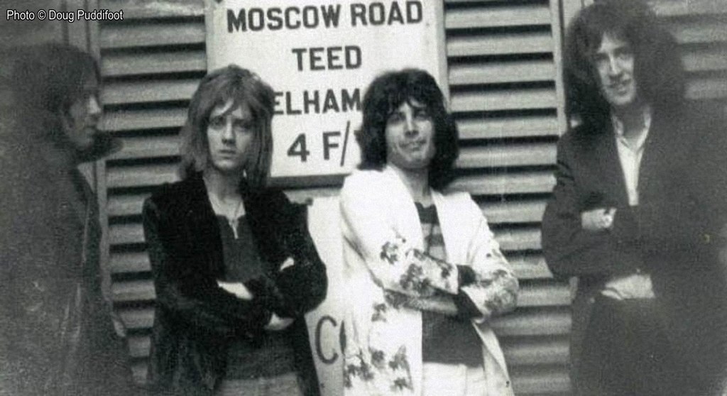 A black and white photo of an early picture of the band members of Queen standing outside a venue. Left to right; Mike Grose, Roger Taylor, Freddie Mercury and Brian May.