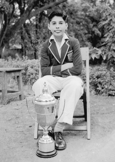 A black and white photo of Freddie Mercury at around age eight. He sits on a chair smiling, wearing a school uniform with a trohpy next to his feet.