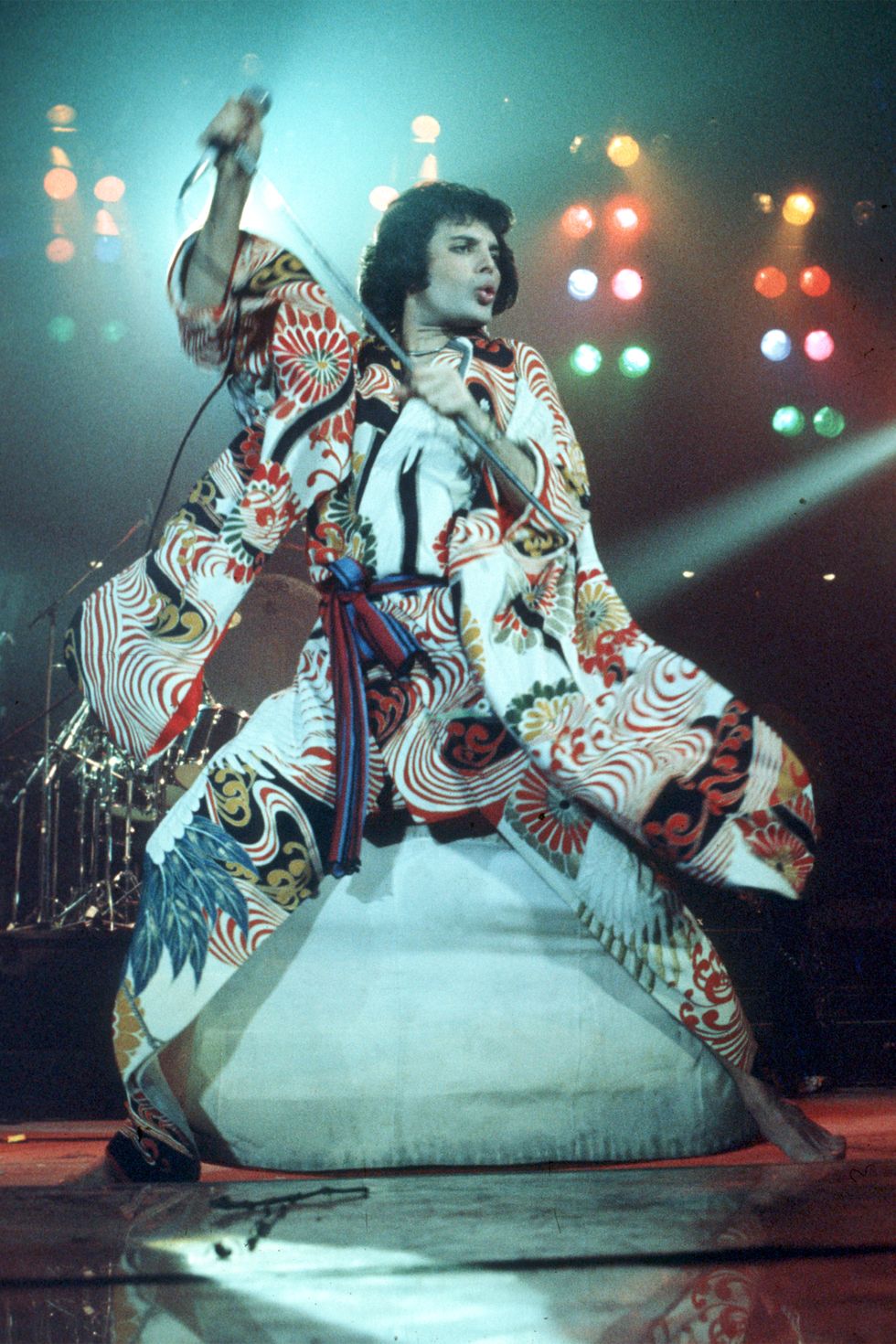 a picture of Freddie Mercury wearing a dazzling multicolor coat while he preforms on stage under bright lights