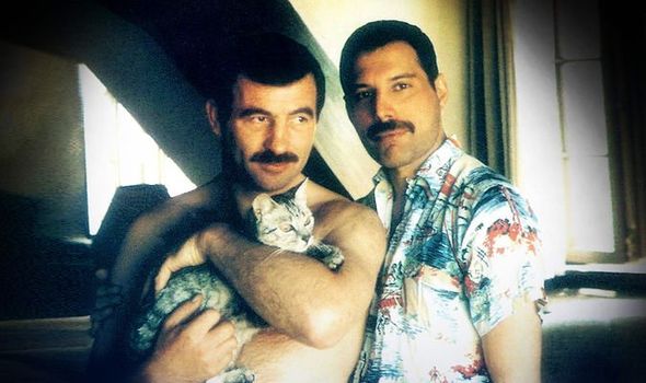 A picture of Freddie Mercury and his partner Jim Hutton. They stand next to each other at home. Freddie is smiling while Jim is shirtless, holding their cat.