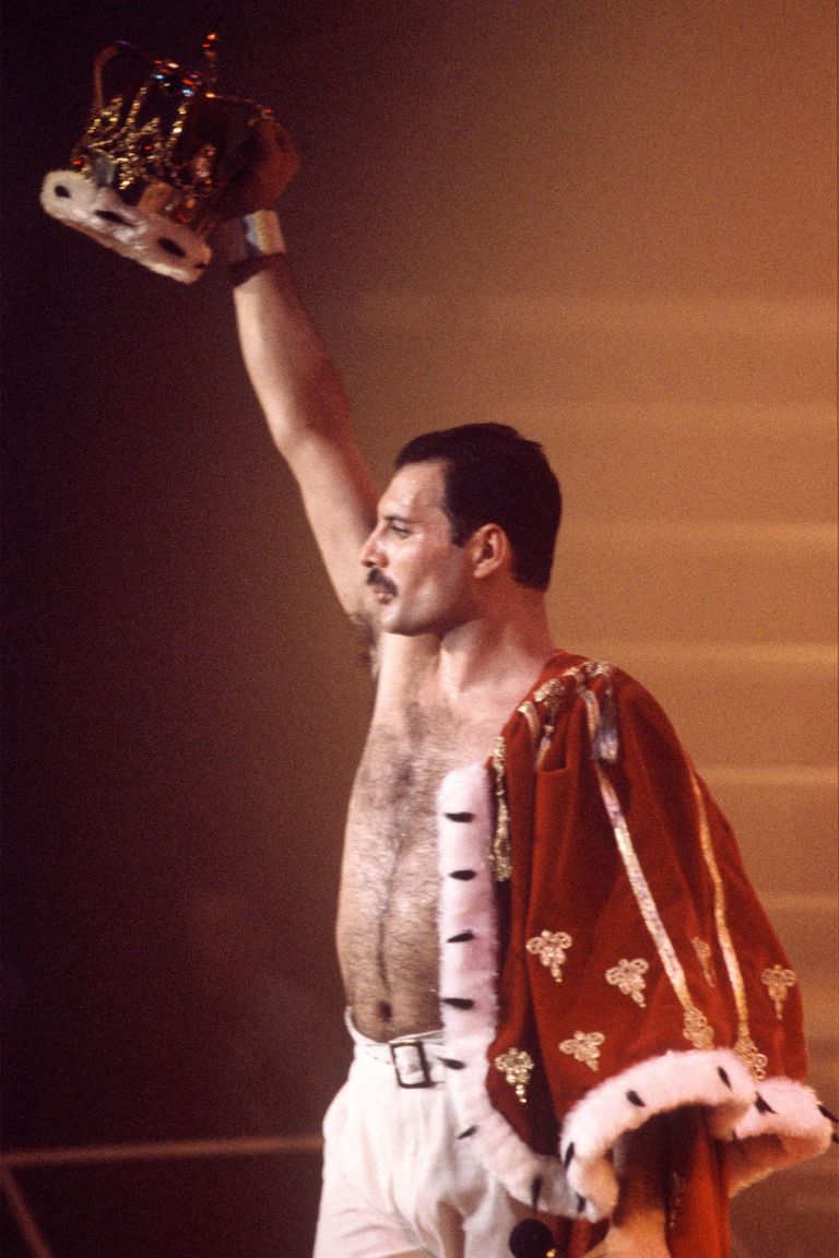 a picture of Freddie mercury wearing a red velvet cape draped on one shoulder and him holding up a royal looking crown while he looks out at an audience