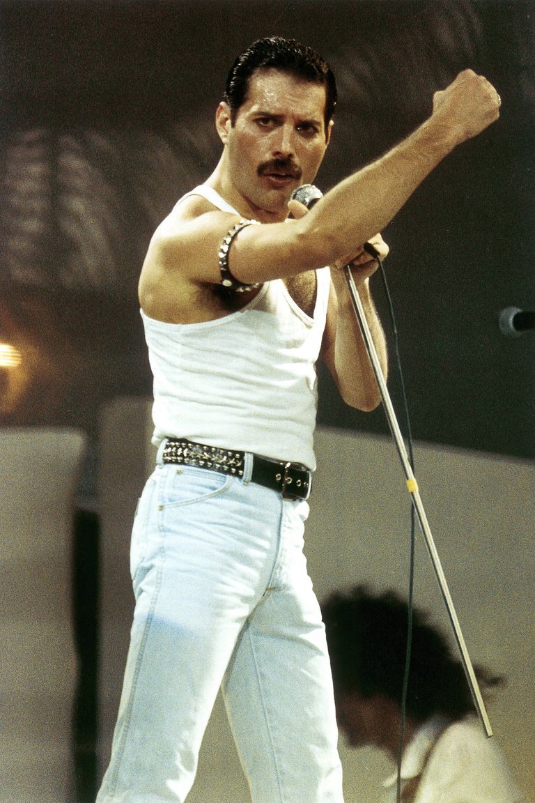 a picture of Freddie Mercury in a white tank top singing into a microphone while he makes as first at a crowd