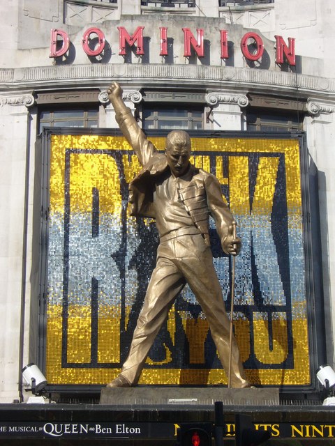 A picture of a bronze statue of Freddie Mercury cast in the likeness of his yellow jacket outfit, with one fist in the air and one on a microphone. It stands in front of a large building titled 'Dominion'.