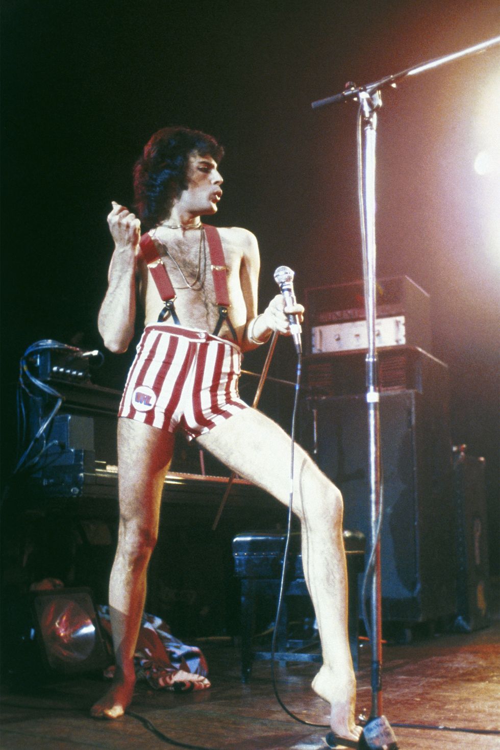 a photo of Freddie Mercury wearing red suspenders and tight striped red and white shorts striking a dramatic pose while he preforms on stage