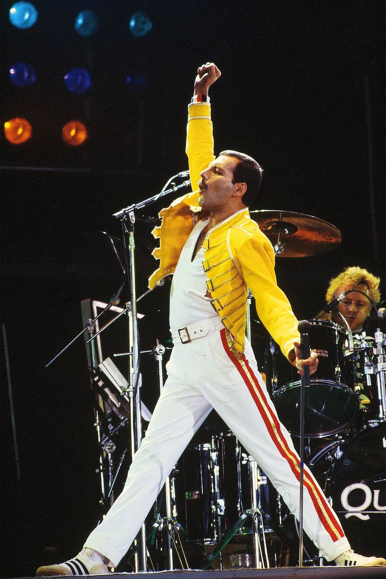 a picture of Freddie Mercury preforming on stage in white pants and a bright yellow jacket while he pumps on first into the air