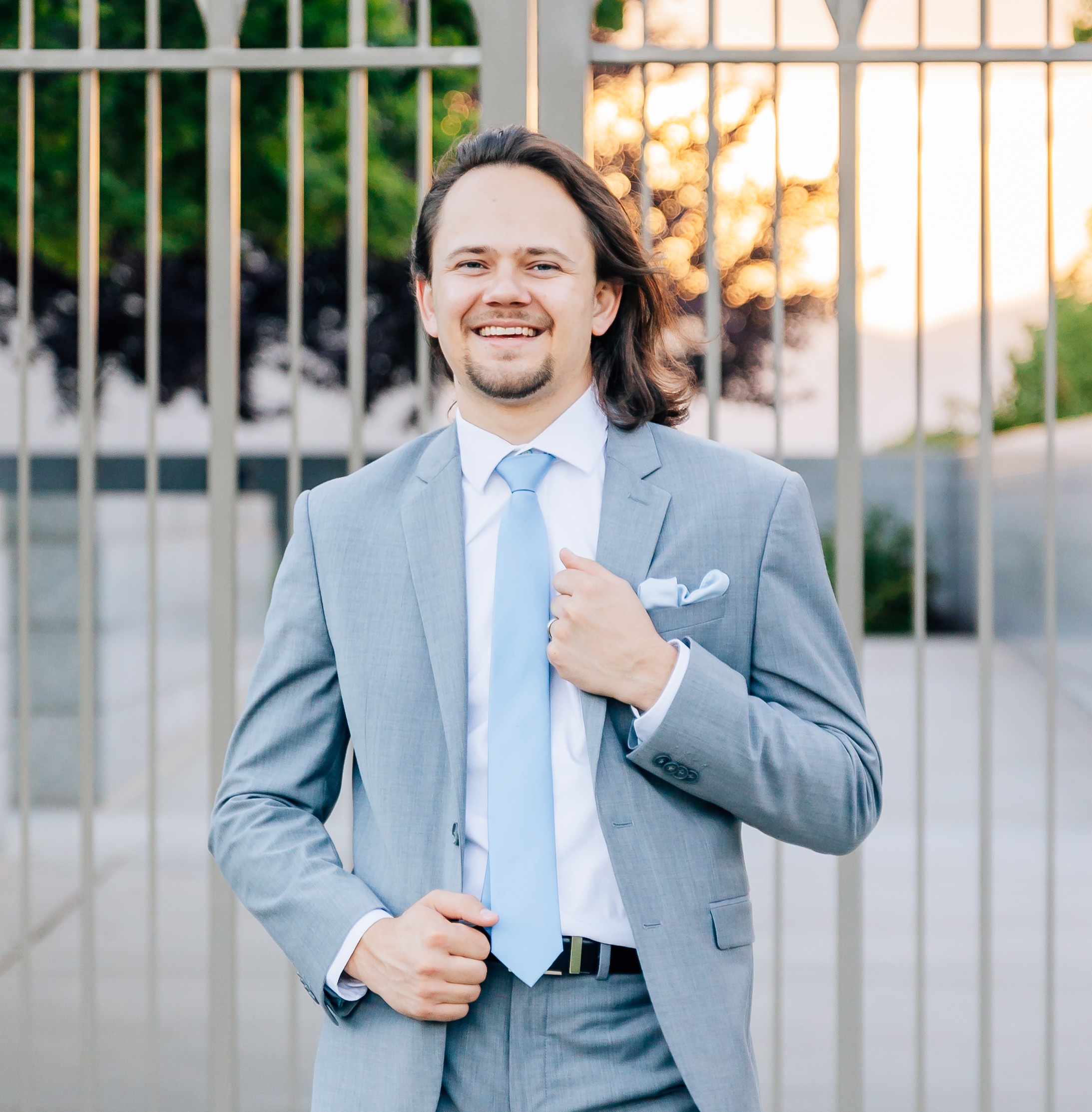 A dashing young man with long hair blowing in the wind posing in front of a gate wearing a grey suit with a white shirt and dusty blue tie