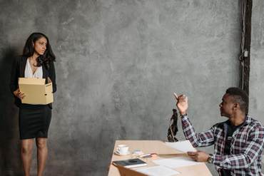 An image of woman stands off in a corner seeming nervous while a man sitting at a desk points at her saying something unknown.