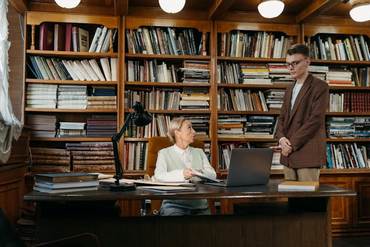 A picture of an older woman sitting at a desk surrounded by bookcases showing something to a young man who stands respectfully off to the side listening.