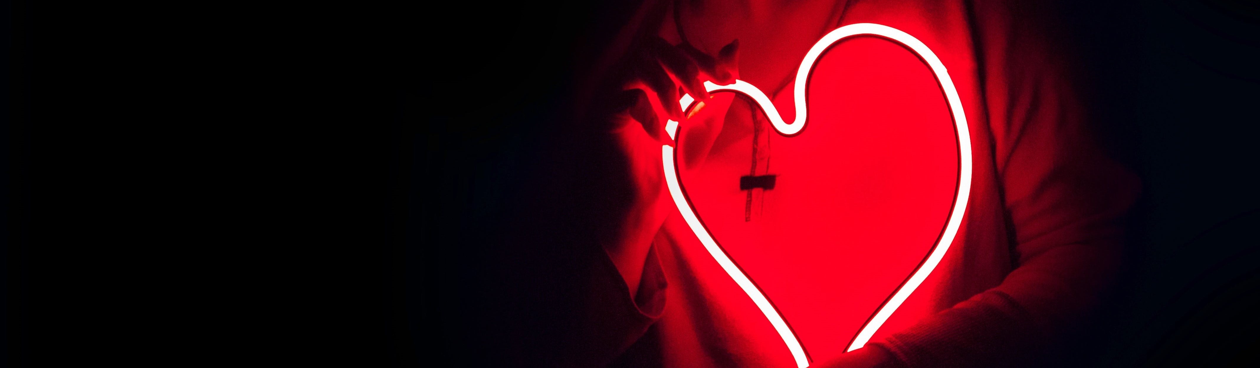 banner image of a person in the dark holding a heart shaped LED light at chest level. The light gives off a red glow.