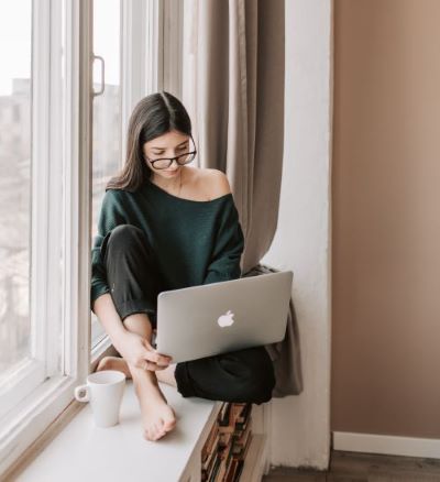 an image of a woman sitting on a windowill with a mug next to her, she is holding a laptop on her thigh and looking down at it